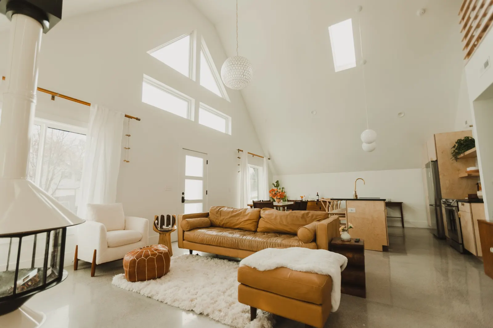 Bright, spacious living room with a high, vaulted ceiling and multiple skylights. Features a tan leather sofa, a white armchair, a round leather pouf, and a cream-colored shag rug. The room has large windows with white curtains and a modern fireplace in the corner.