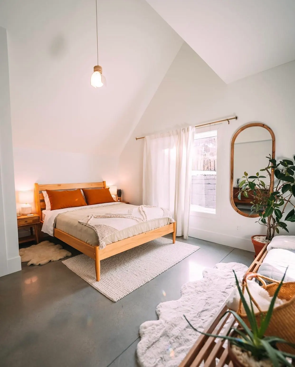 A cozy bedroom with a wooden bed, white bedding, and rust-colored pillows. The room has white curtains, a large mirror with a wooden frame, and a potted plant. The floor is concrete with rugs, and there's a hanging light fixture on the ceiling.