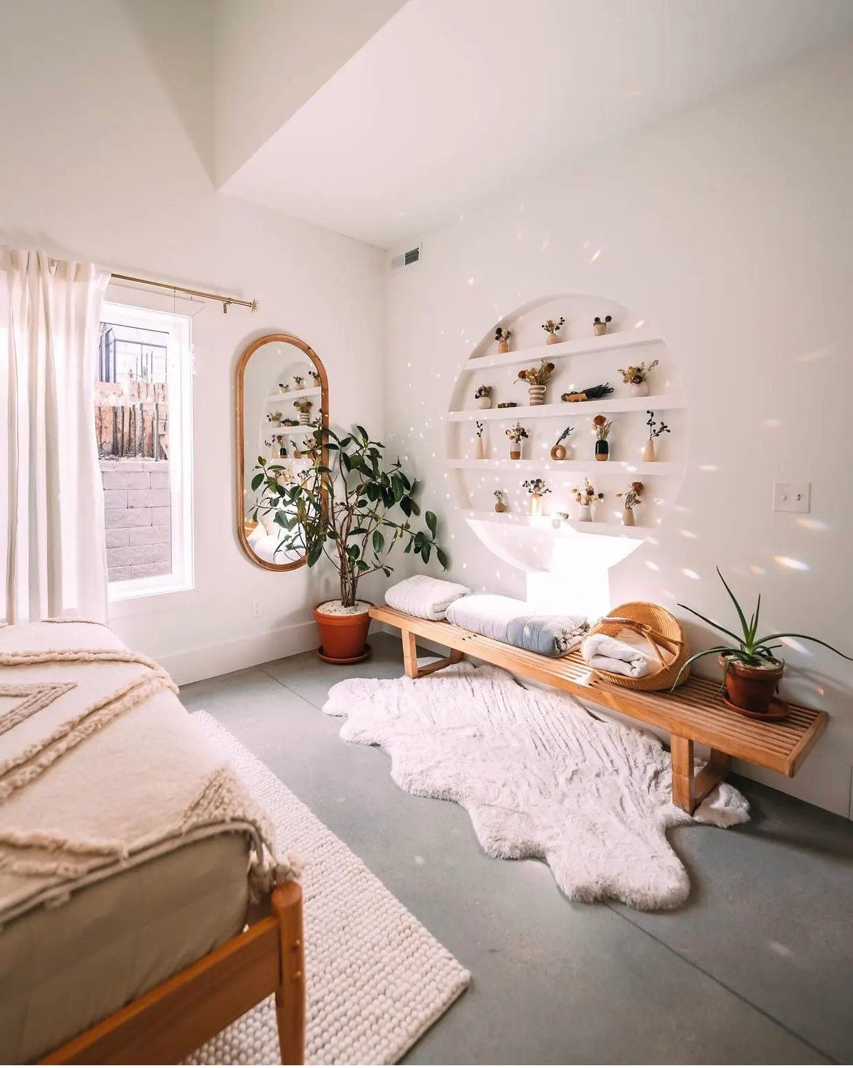 Bright, minimalist bedroom with a large window, white walls, a wooden bench with rolled towels, indoor plants, a white rug, and decorative shelves with small vases and flowers.