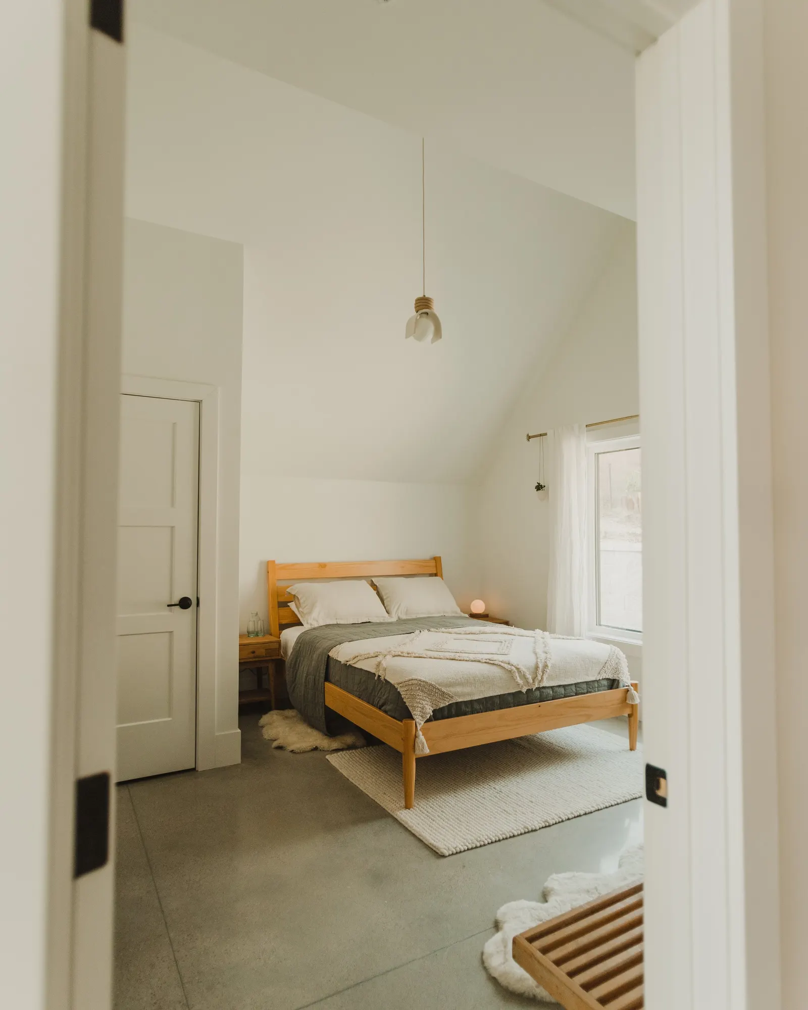 A cozy bedroom with a wooden bed frame, light bedding, a small nightstand with a lamp, and a window with white curtains, all in a minimalist style.