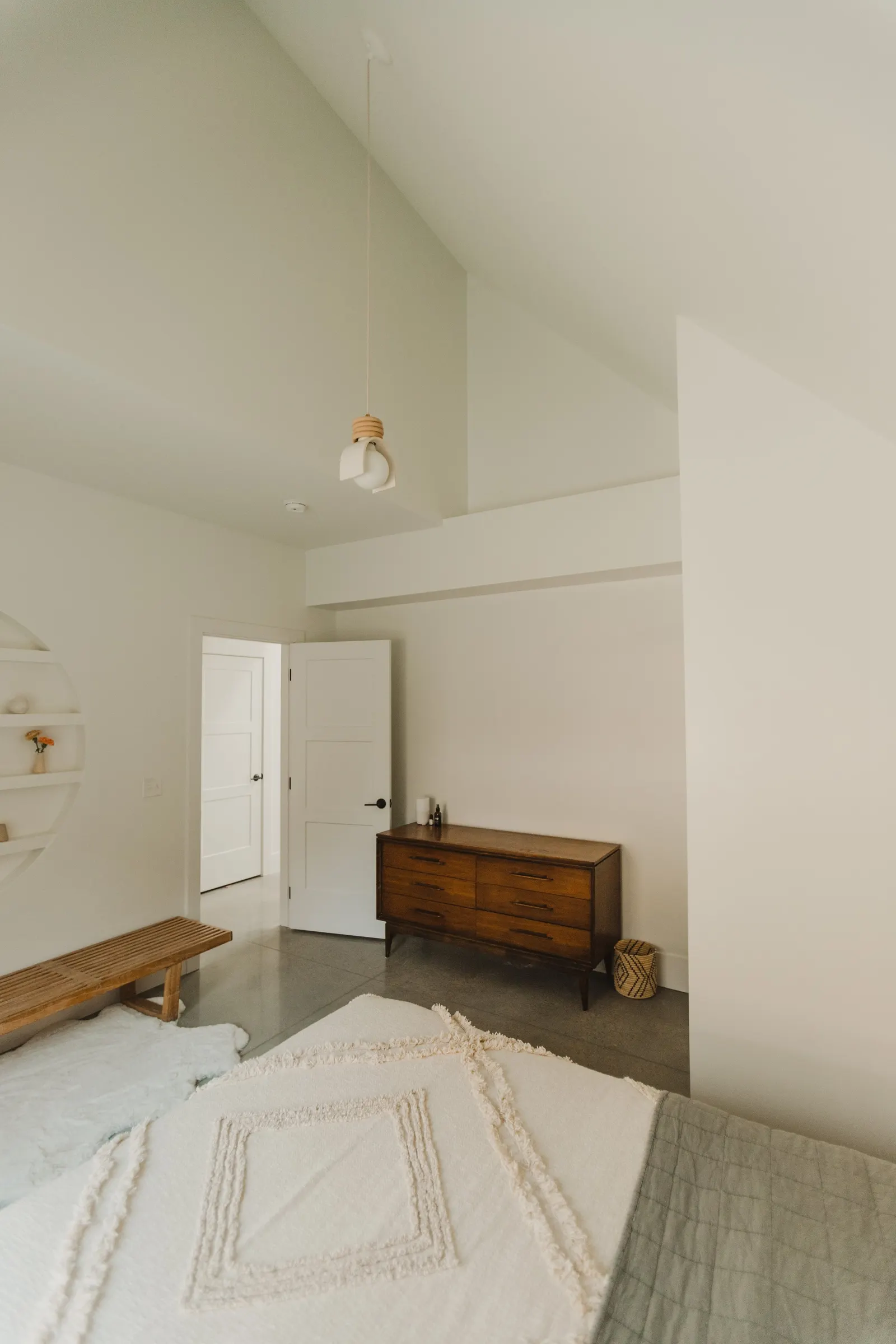 A bedroom with a white wall, a wooden dresser, a bench, and a circular wall shelf with small decor items. There is an open door and a ceiling light fixture.