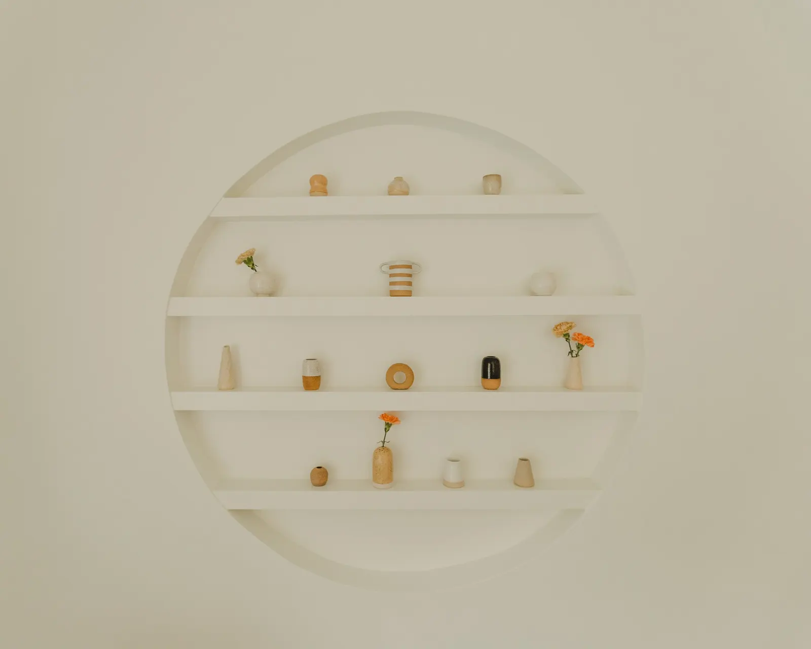 White circular wall shelf with multiple small vases and decorative items, some with flowers, on a plain wall.