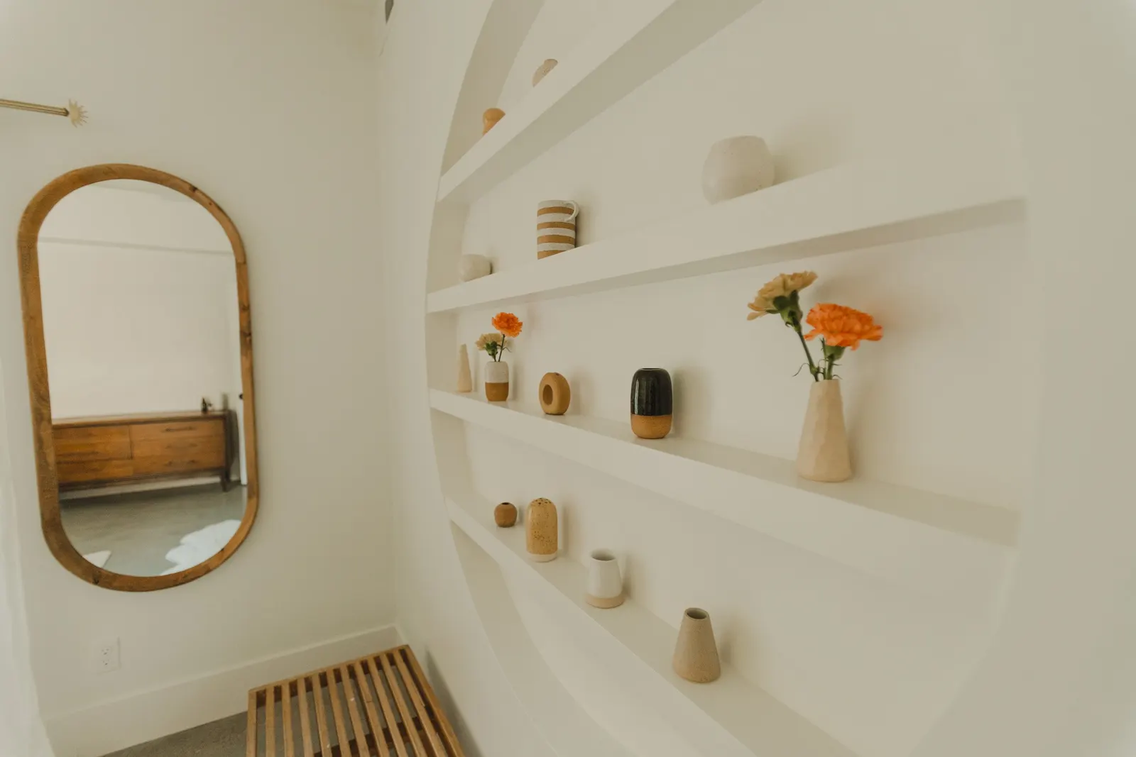 White wall with built-in curved shelves holding small vases and decorative objects, with a wooden slatted bench and a wooden-framed oval mirror reflecting a bedroom with a wooden dresser.