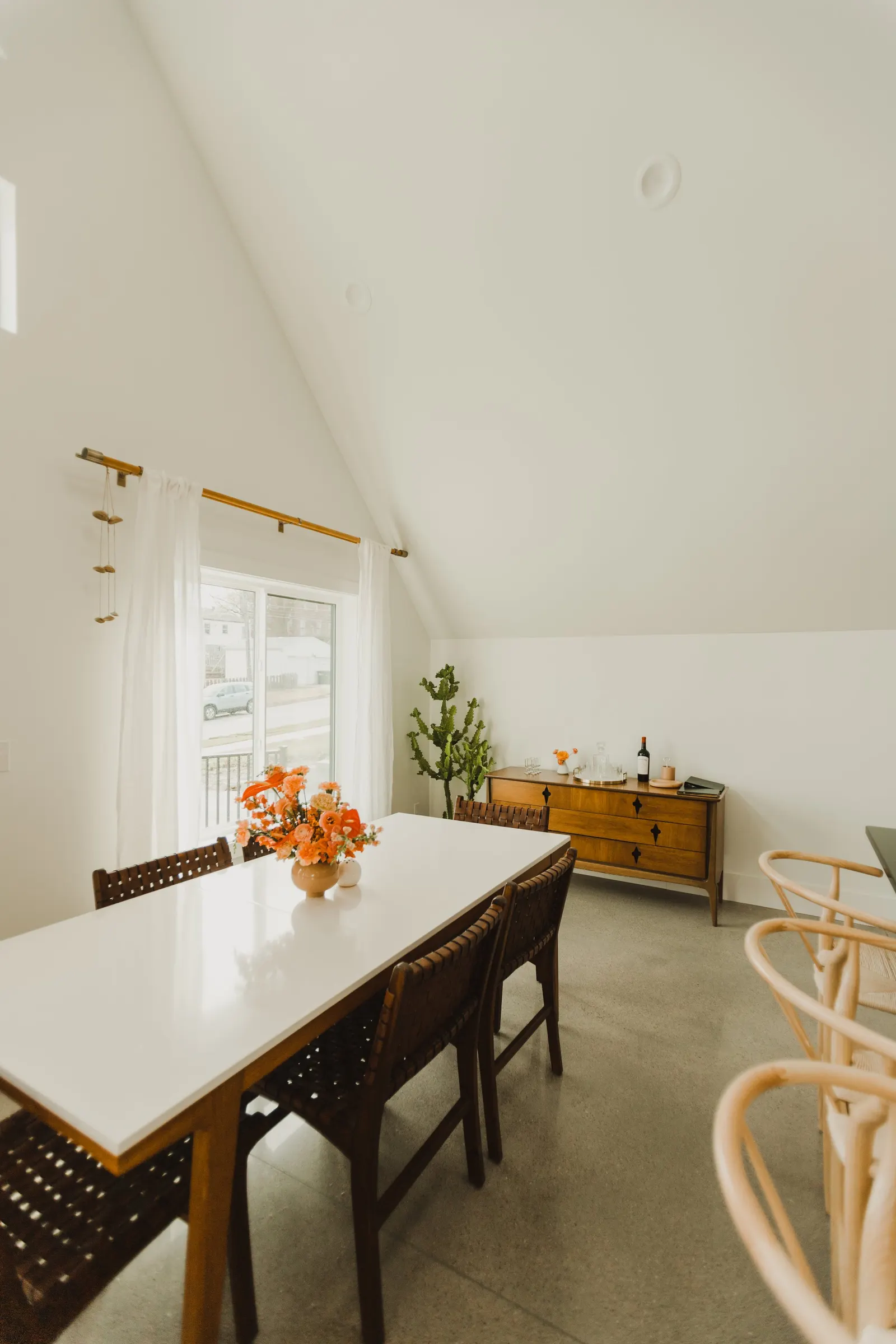 A room with a white ceiling slanted on the right side, cream-colored walls, a window with white curtains, a white dining table with a flower arrangement, four wooden chairs, a wooden sideboard with bottles and glassware, a cactus plant, and a small h