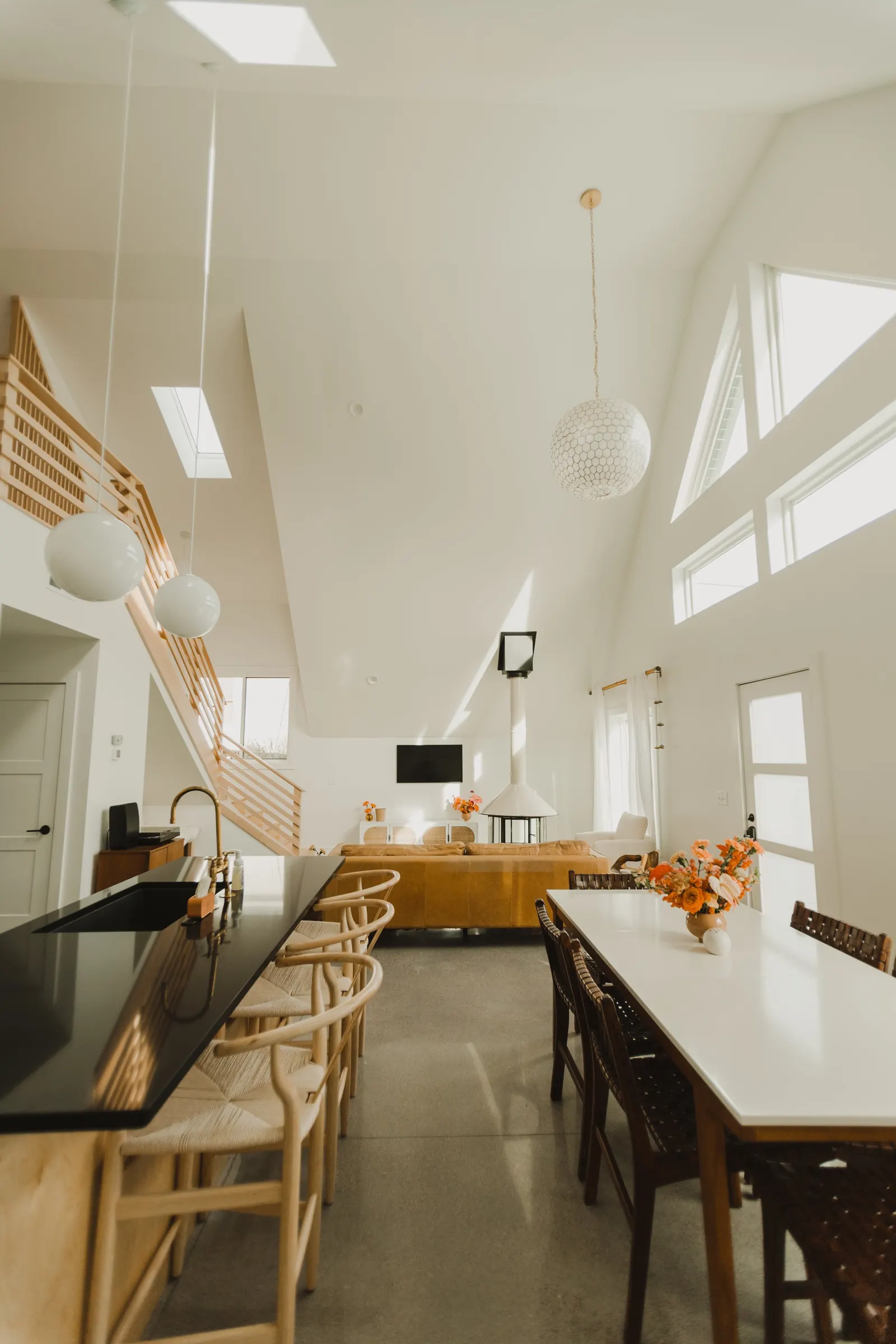 Open-concept living and dining area with high vaulted ceiling, large windows, and modern decor. There is a kitchen island with beige bar stools, a main dining table with chairs, a sofa, a flat-screen TV, and a hanging pendant light. Bright natural li