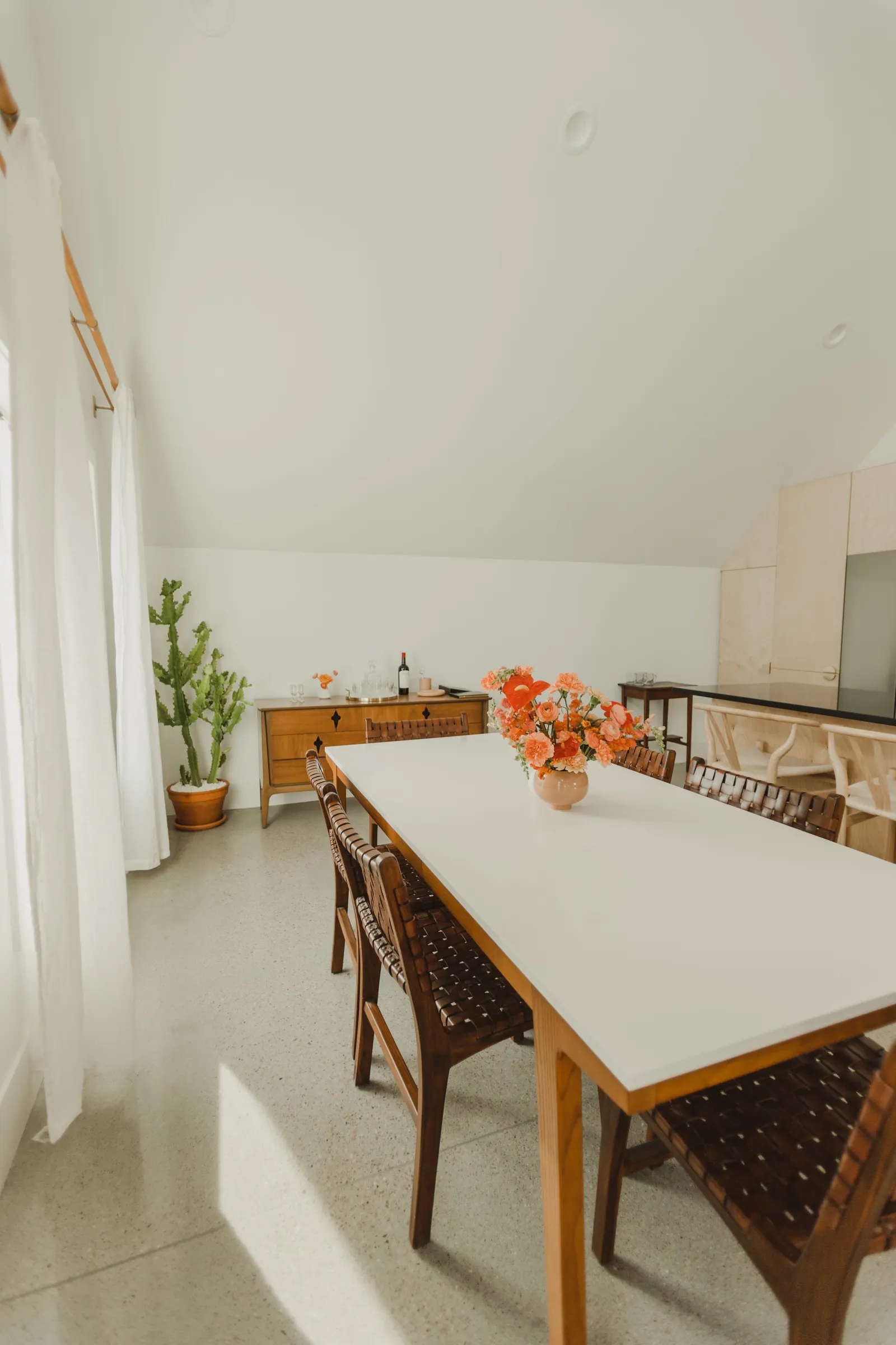 A dining room with a white table, brown woven chairs, a large bouquet of pink and orange flowers, a wooden sideboard with a bottle of wine and glassware, a large cactus, and a window with white curtains.