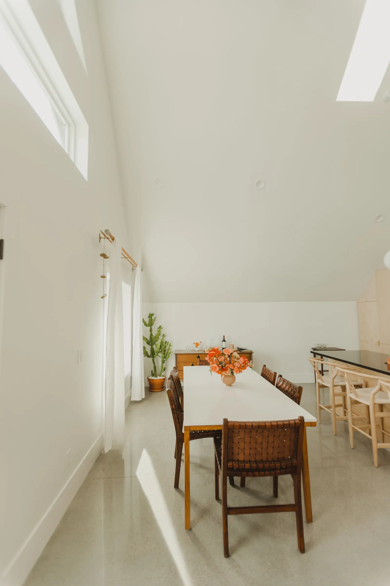 Dining room with a rectangular white table, surrounded by six woven chairs, a vase with orange flowers on the table, a green cactus in a pot in the corner, and a sideboard with a wine bottle and glasses, with windows allowing natural light.