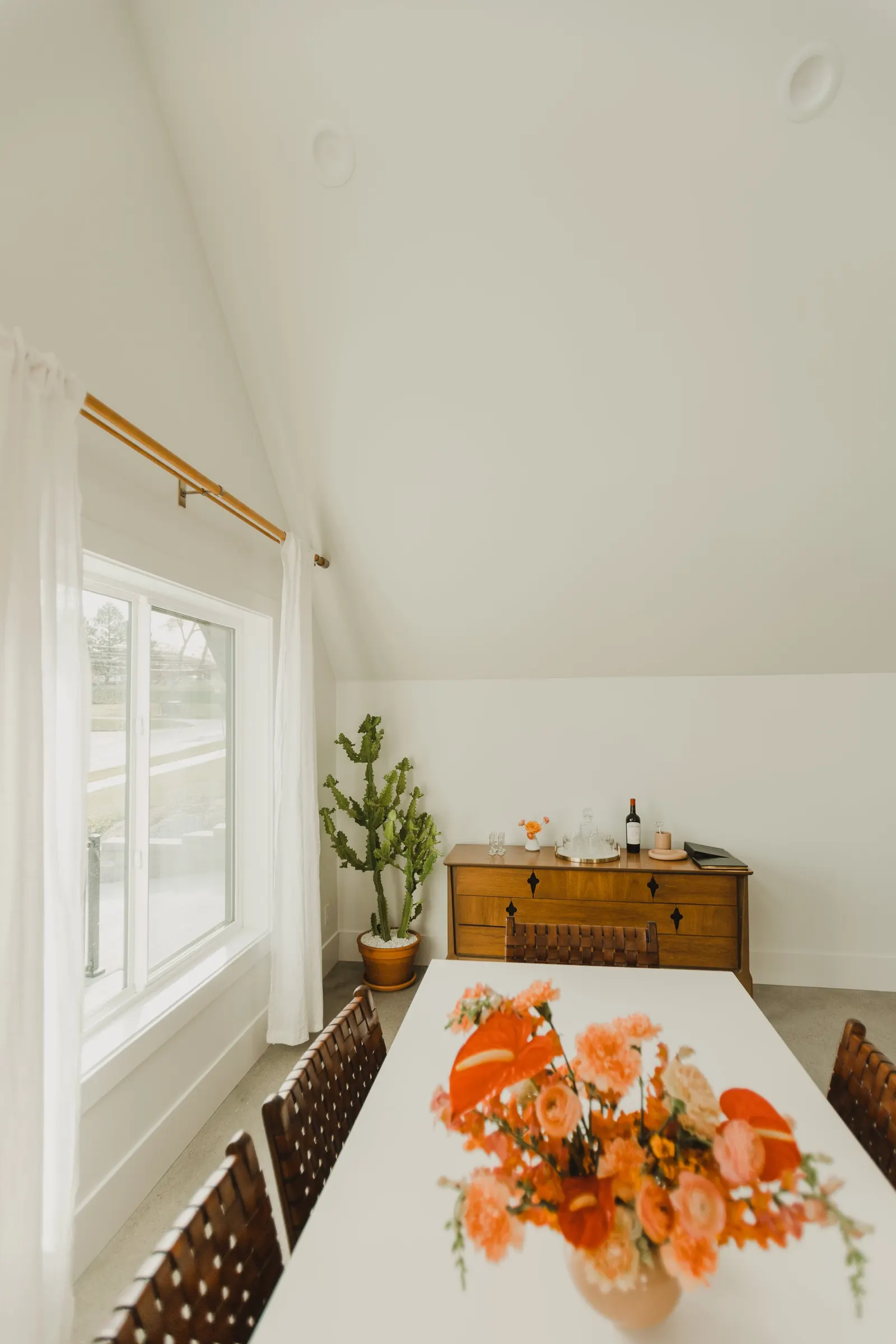 Dining room with a white table, floral arrangement, wooden sideboard, potted cactus, window with white curtains, and various decorative items.