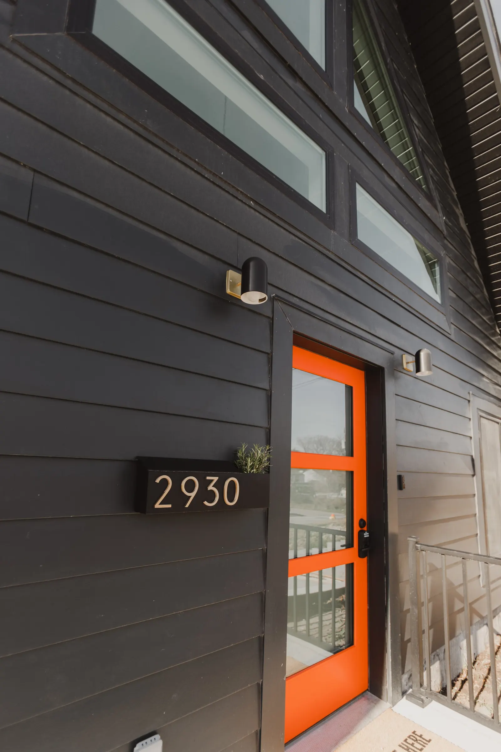 Black wooden exterior of a building with a bright orange door and the number 2930 on a black mailbox with a small plant inside. There are two modern wall lights above the door.