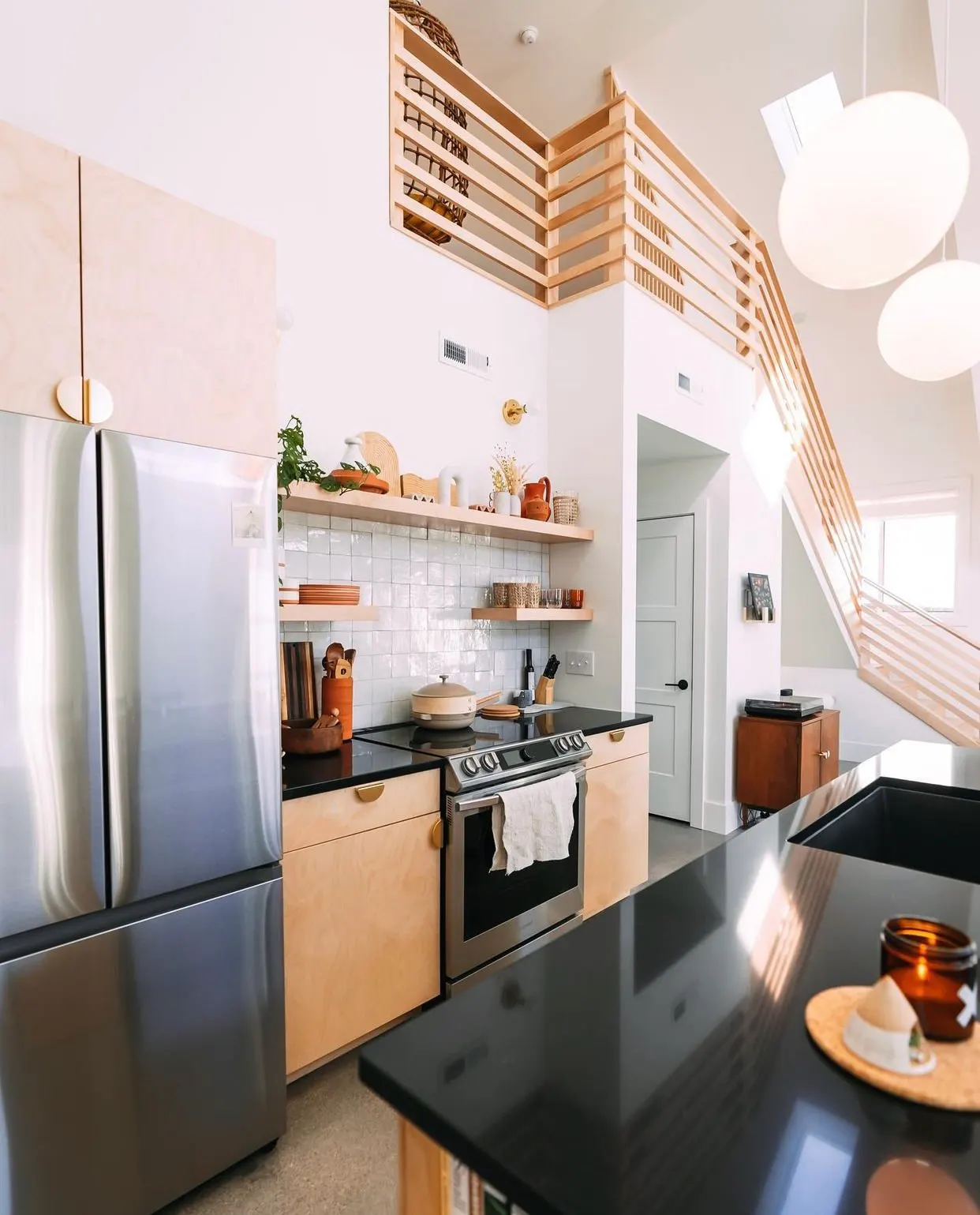 Modern kitchen with light wood cabinets, black countertop, stainless steel refrigerator, and a diagonal wooden staircase leading to a loft area.