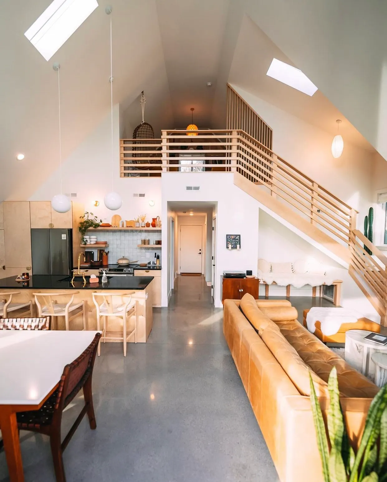 Open-concept living room with a tan leather sofa, a small side table, and houseplants. A kitchen with a black countertop, light wood cabinets, and hanging globe pendant lights is visible, along with a staircase with wooden railings leading to a loft