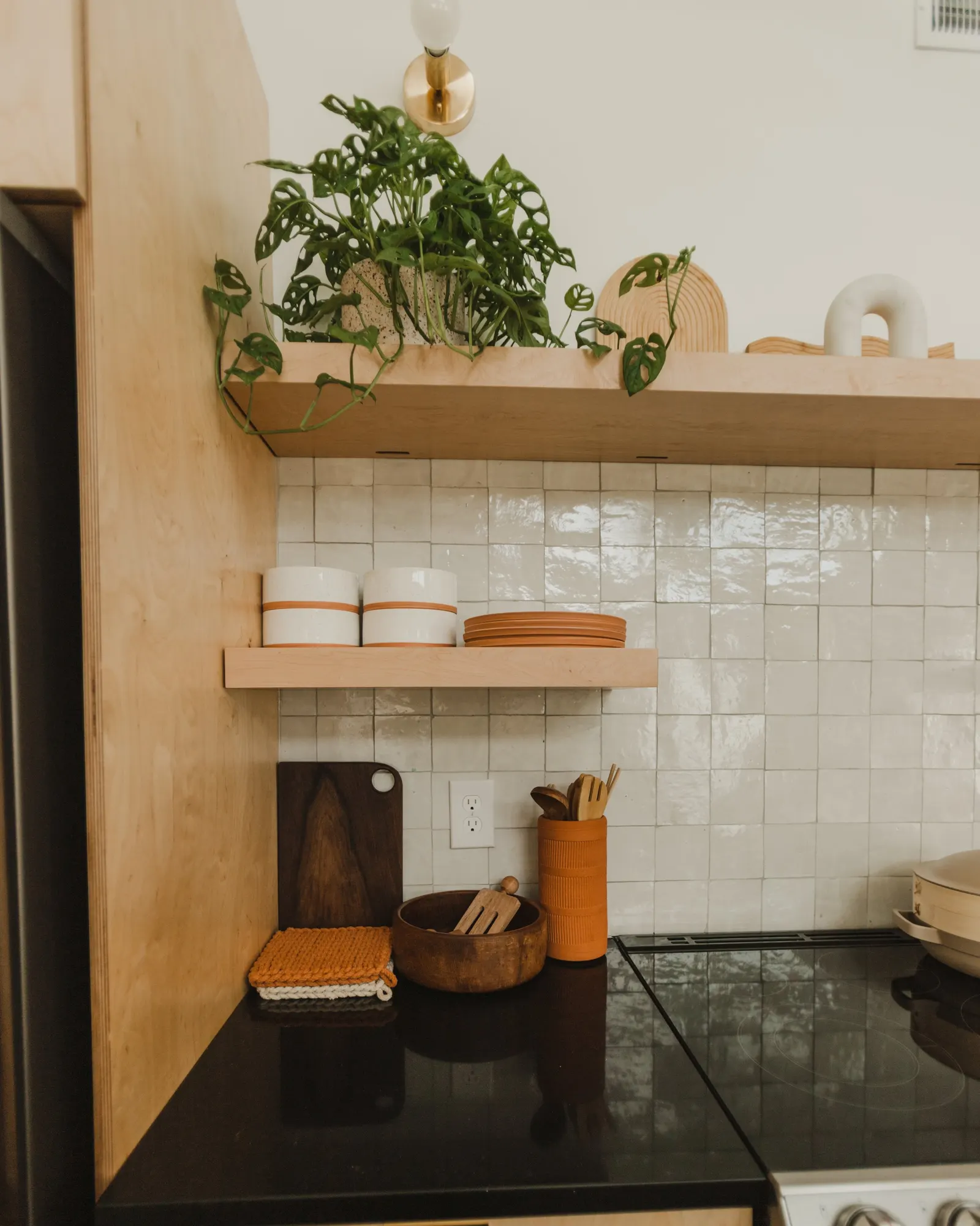 Kitchen countertop with wooden shelves, a potted Monstera plant, ceramic bowls, brown plates, a wooden cutting board, a container holding utensils, a small wooden bowl with a spoon, and a folded colorful dish towel.