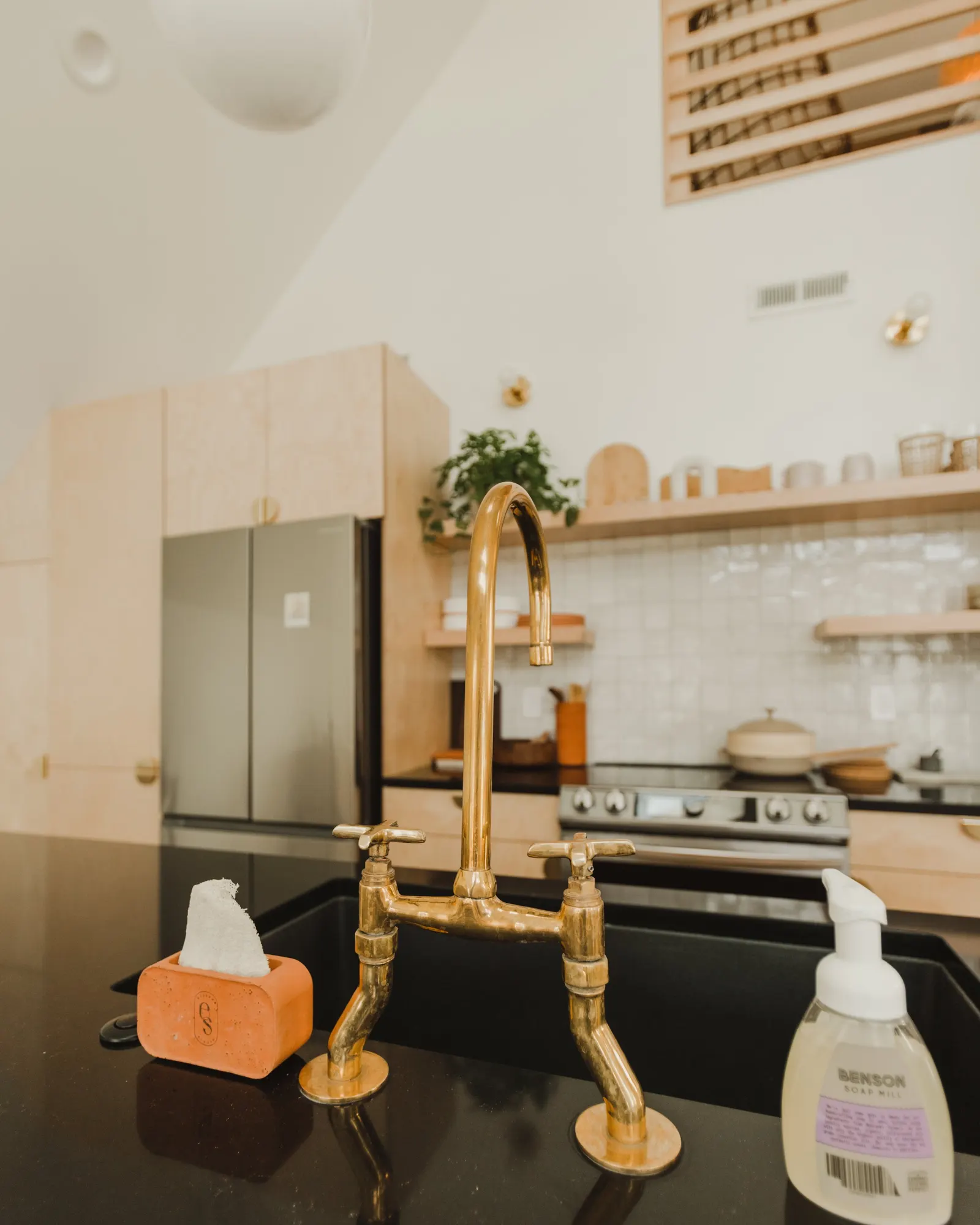 Kitchen countertop with a brass faucet, soap dispenser, and sponge. Background features a modern kitchen with a stove, wooden cabinets, open shelves with dishes, and a potted plant.