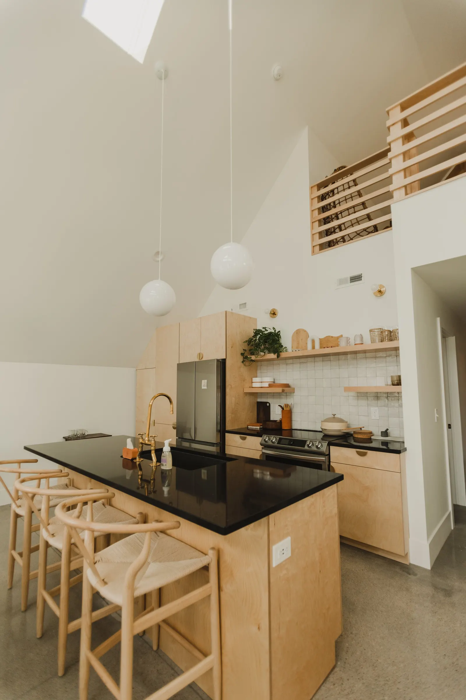 Modern kitchen with light wood cabinets, black countertop island, gold fixtures, white walls, and hanging white spherical pendant lights.