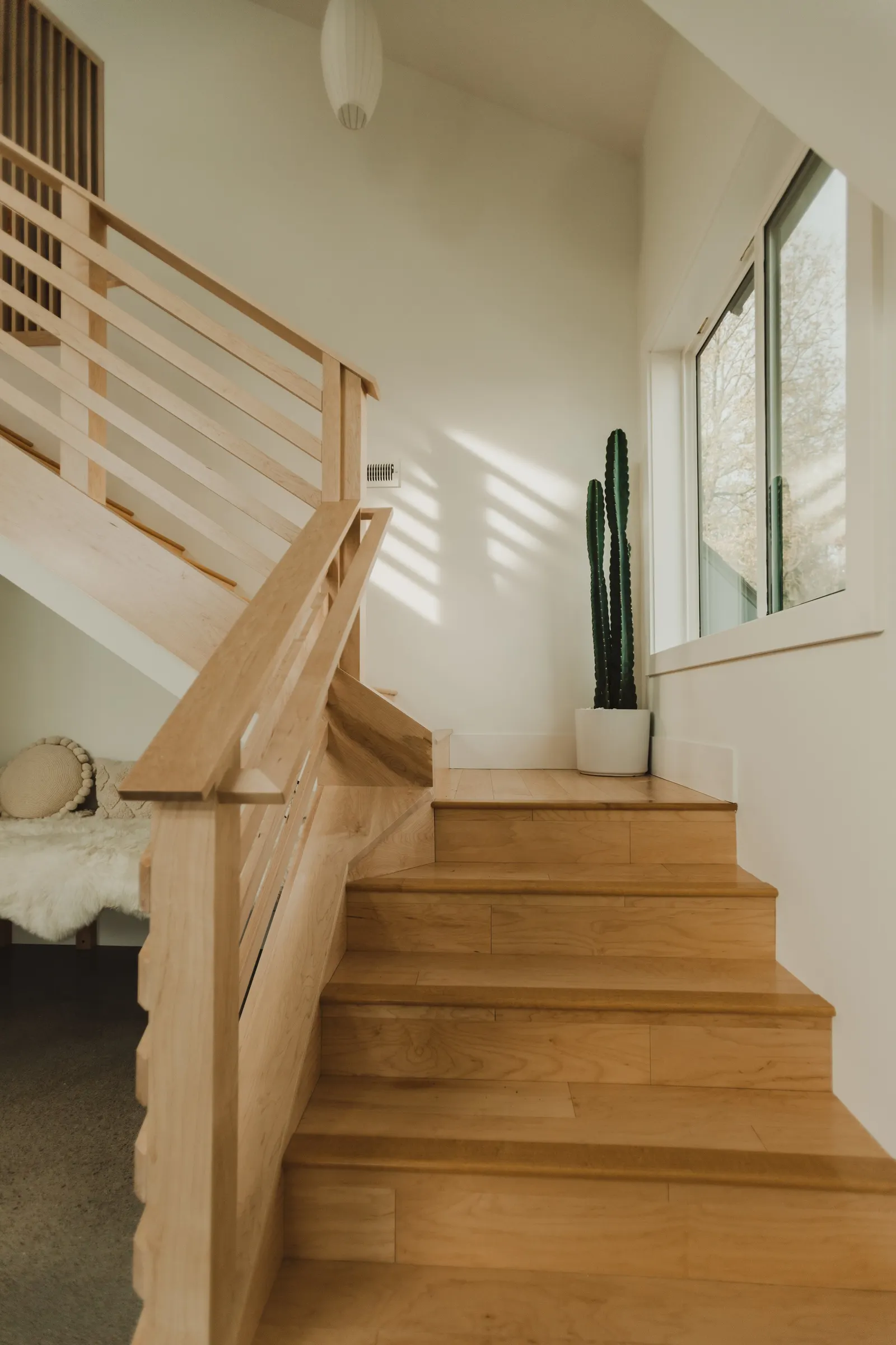 Wooden staircase with railing leading to a bright room with a large window, a potted cactus plant, and a corner seat with a round pillow and stuffed animal.