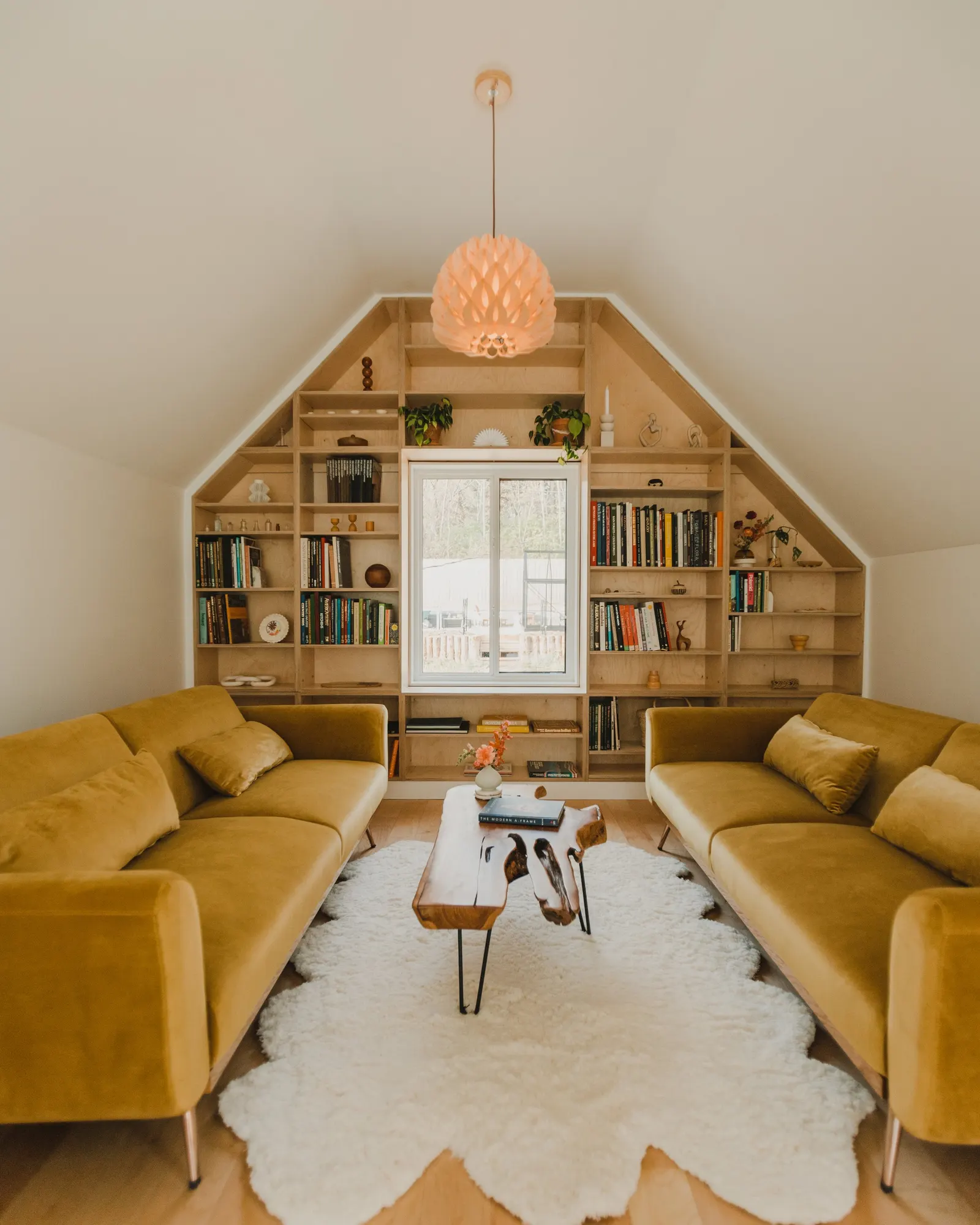 Living room with mustard yellow couches, a white shag rug, a wooden coffee table, and a built-in bookshelf on the wall with a window in the center.
