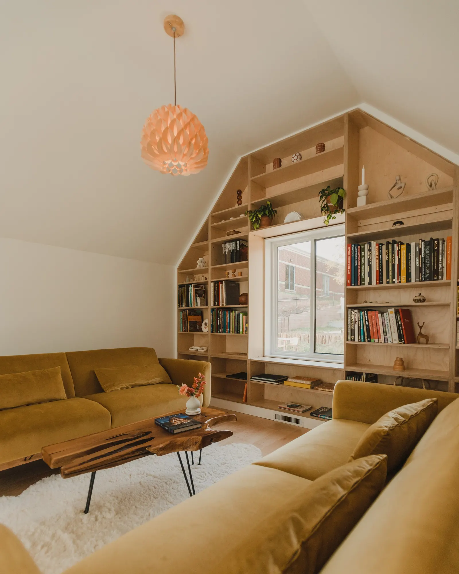 Living room with two mustard-colored sofas, a wooden coffee table with a book and a vase of pink flowers, a window with built-in wooden bookshelf filled with books and decorative items, a soft white rug, and a pink pendant light hanging from the ceil