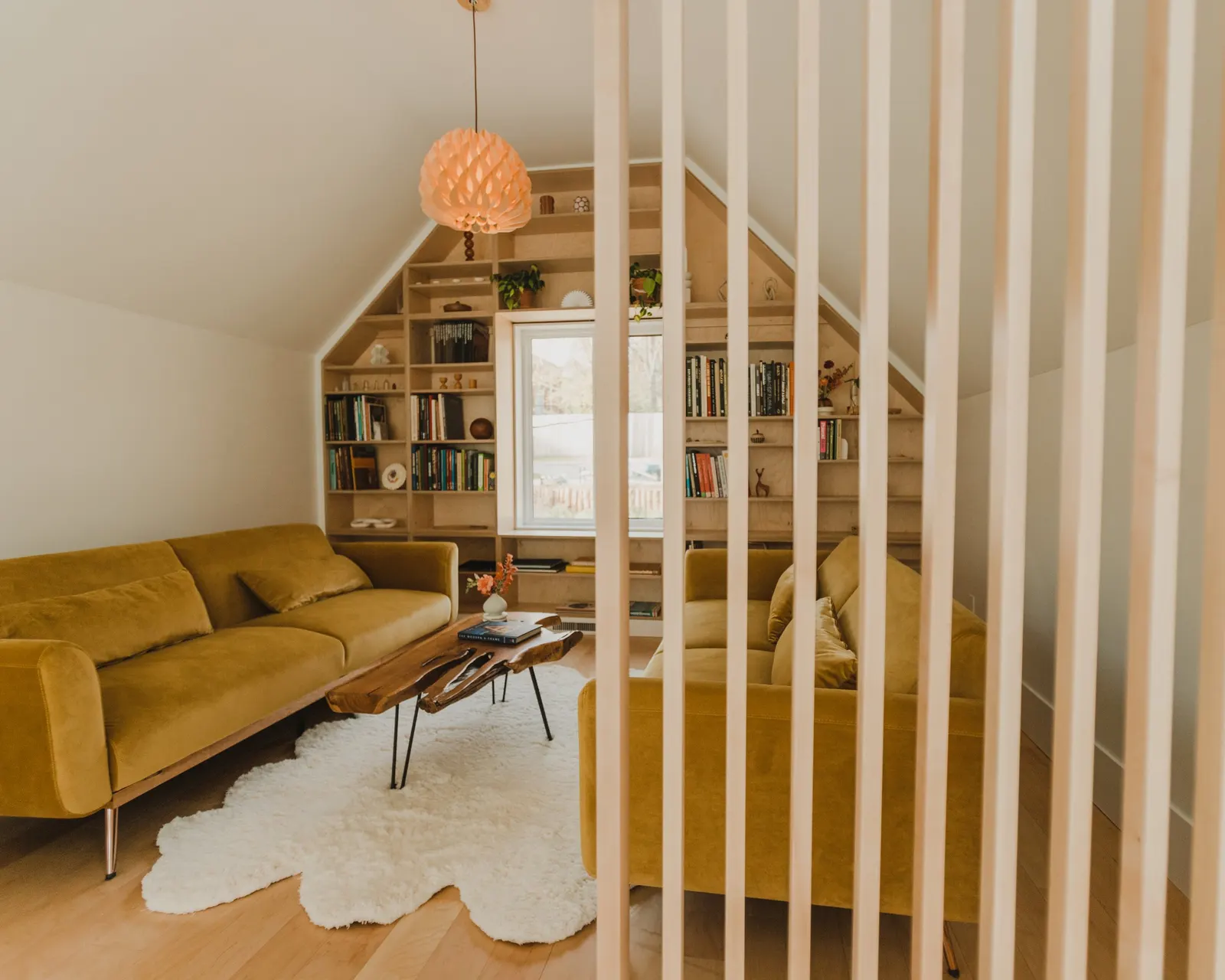 Living room with yellow sofas, a wooden coffee table, a white fluffy rug, built-in bookshelf under a staircase, and a window with views outside.