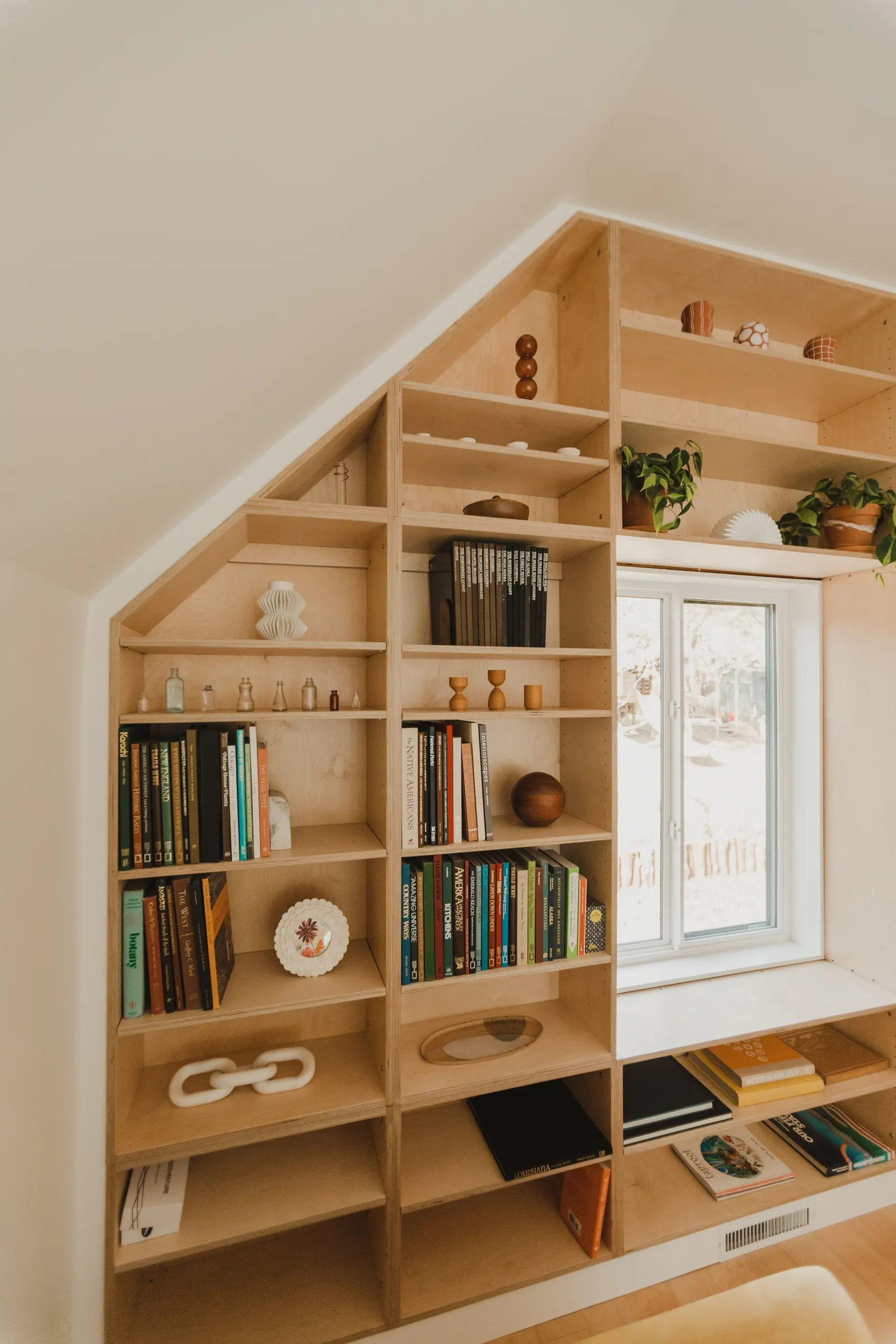Wooden bookshelf with eclectic decorations and books, built into a wall with a sloped ceiling and a window
