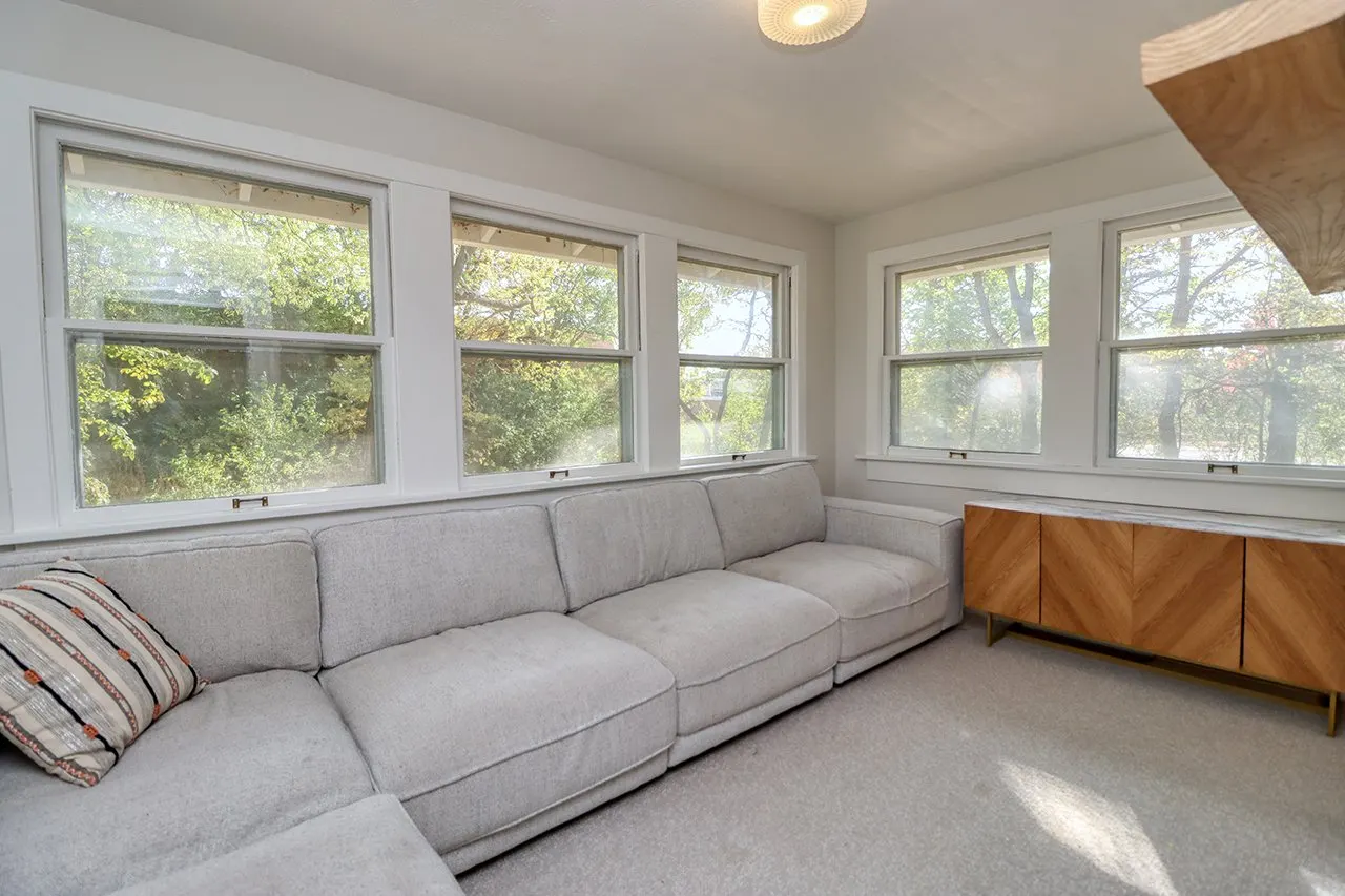Living room with a beige sectional sofa, a patterned throw pillow, and four large windows showing trees outside. There is a wooden cabinet against the wall and a ceiling light fixture.