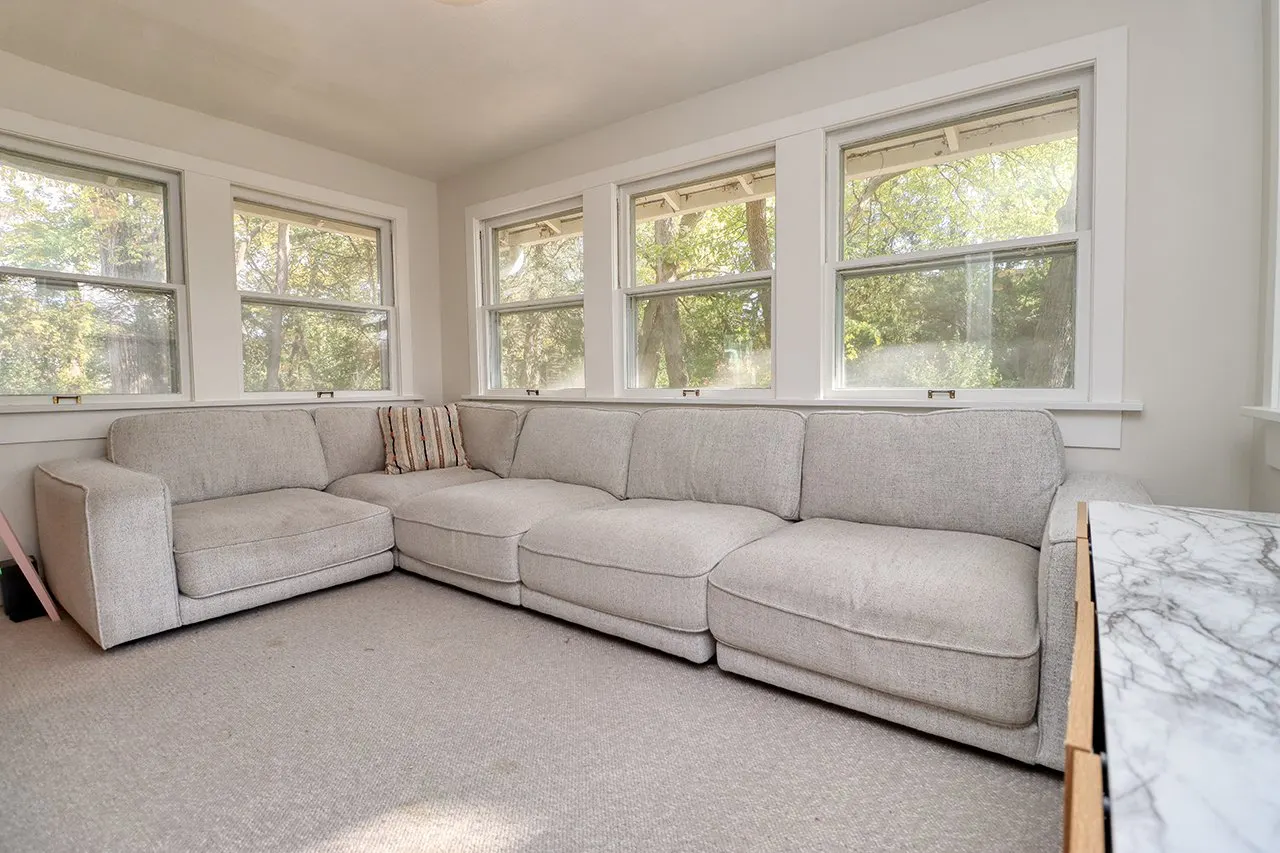 Living room with a beige sectional sofa in front of a wall of five large windows showing trees outside.