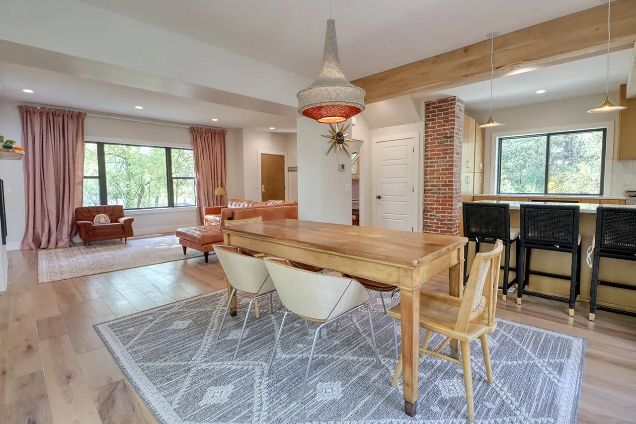 Open-concept living and dining area with large windows, pink curtains, hardwood floors, a wooden dining table with six chairs, a brick accent wall, a kitchen with bar stools, and modern light fixtures.