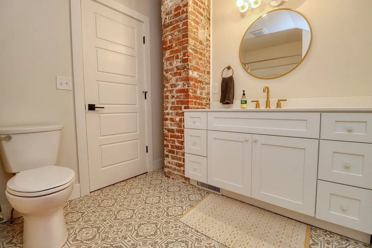 Bathroom with white vanity, round mirror, brass fixtures, brown hand towel, soap dispenser, exposed brick wall, white door, patterned tile floor, and a beige rug.