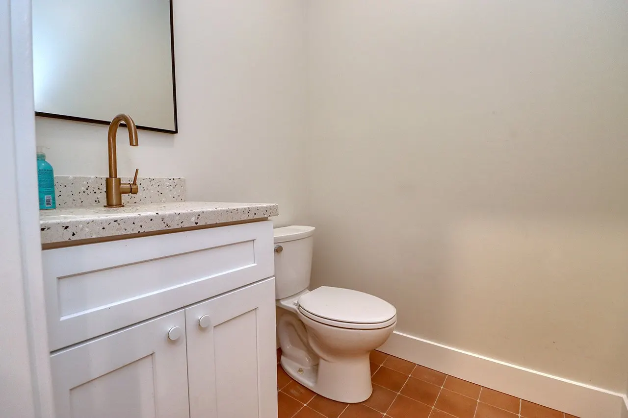 Bathroom with a white vanity, speckled countertop, gold faucet, mirror, and a white toilet against plain white and beige walls with brown tile flooring.
