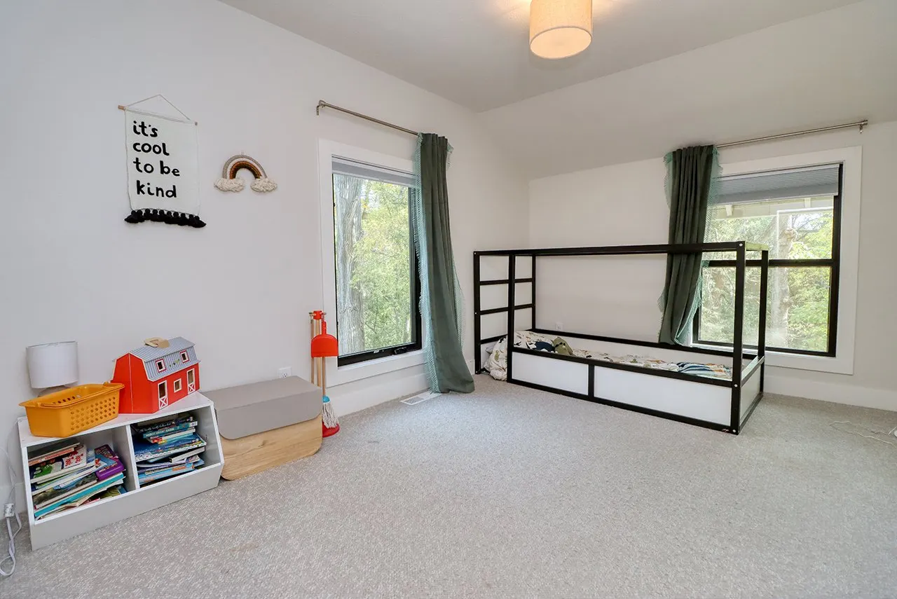 A children's bedroom with two large windows, green curtains, a black bunk bed with a mattress, a bookshelf with books, a toy barn, a lamp, and wall decorations including a rainbow and a banner that says 'it's cool to be kind'.