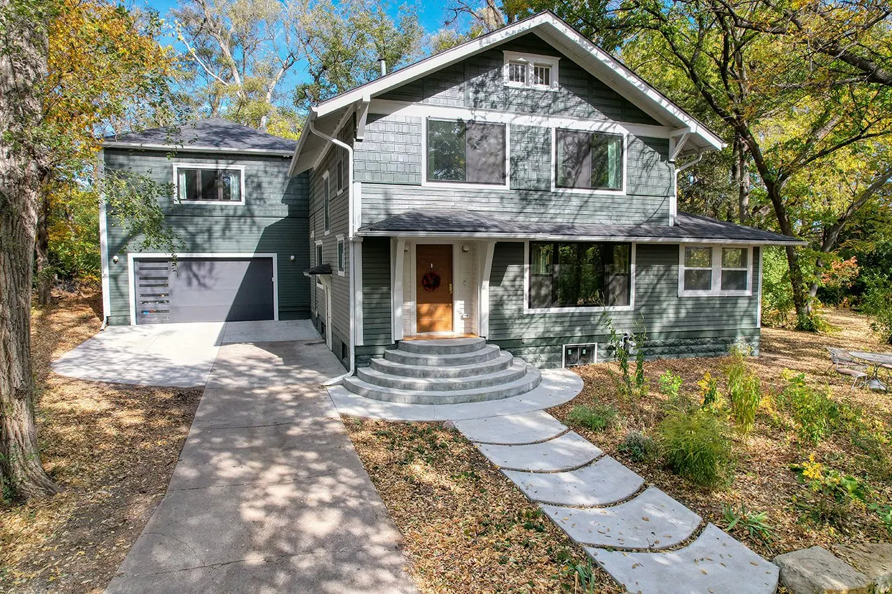 Front view of a modern two-story house with gray siding, surrounded by trees with autumn leaves, a concrete driveway, and a curved walkway leading to the front door decorated with a holiday wreath.