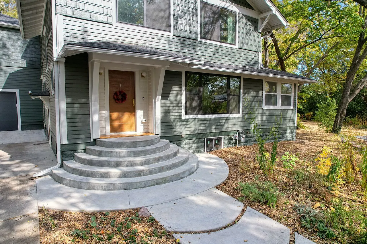 Front view of a two-story house with green siding, a wooden front door decorated with a wreath, and concrete steps leading to the entrance. There are trees and plants around the house on a sunny day.