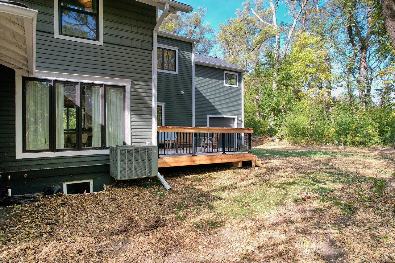 Backyard of a house with a small wooden deck, large windows, an air conditioning unit, and surrounded by trees with a mix of green and fall-colored leaves.