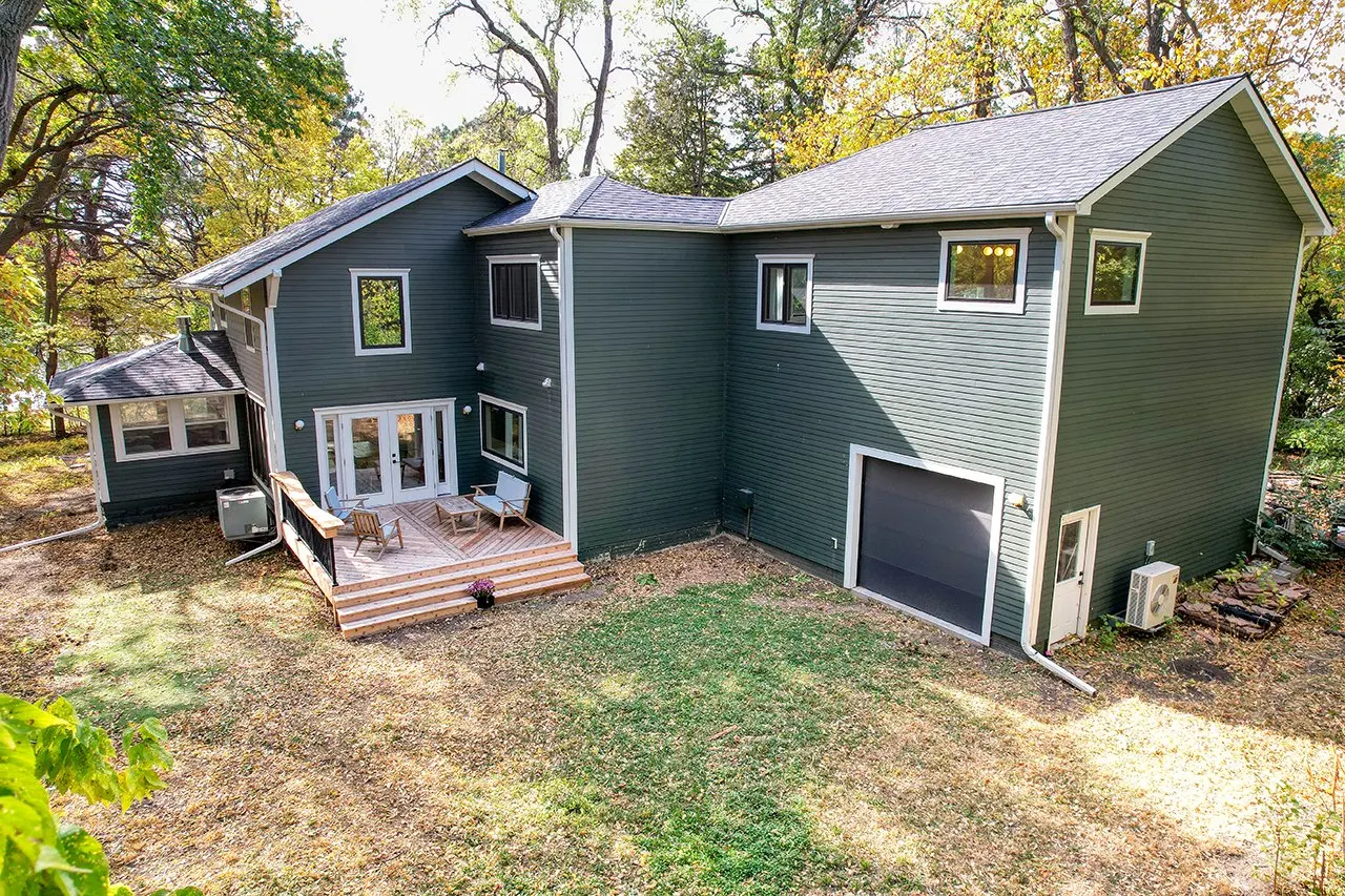 Backyard of a modern two-story house with dark green siding, multiple windows, and a small wooden deck with outdoor furniture, surrounded by trees.