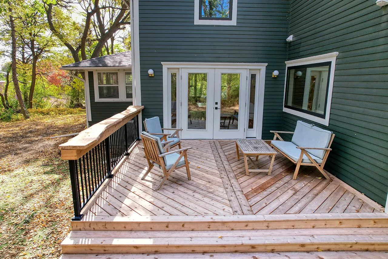 Wooden deck with outdoor furniture, including chairs and a table, attached to a house with green siding and large windows, surrounded by trees.