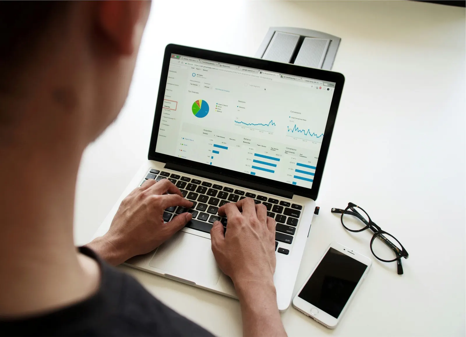 Person analyzing data on a laptop screen, with a smartphone and glasses on a white desk.