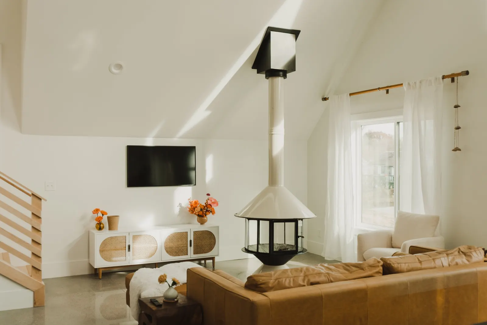 Bright living room with a white ceiling and walls, a flat-screen TV on the wall, a white sideboard with orange flowers, a tan leather sofa, a white armchair near a window with white curtains, and a modern cone-shaped fireplace with a chimney.