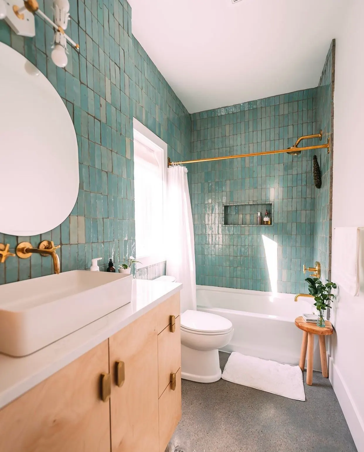 modern bathroom with turquoise tile walls, white bathtub, wooden vanity, white sink, gold fixtures, and a small wooden stool with a plant on it.