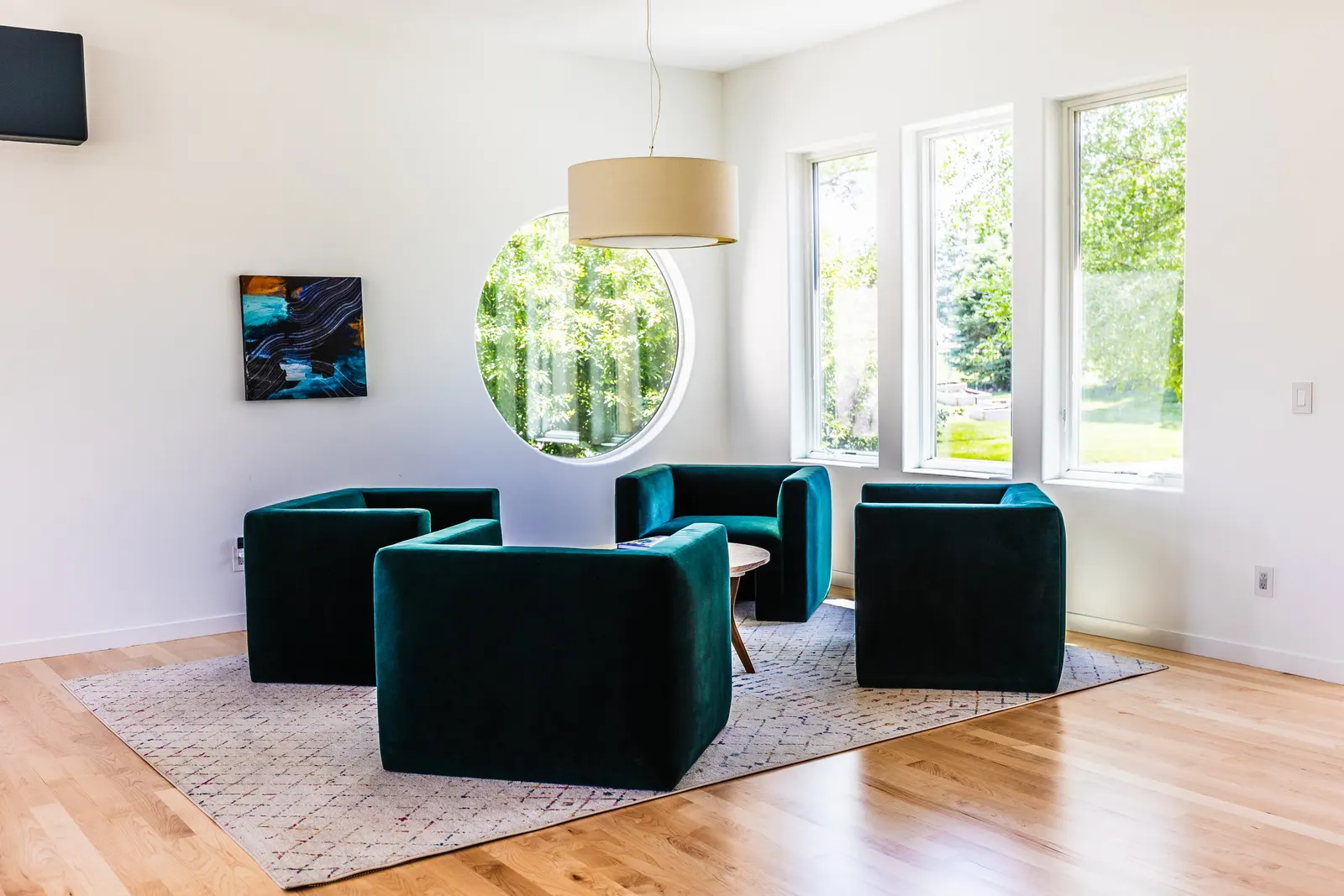 A bright modern room with four dark green armchairs arranged in a circle on a patterned rug, large windows, and a white wall with artwork and a round mirror.