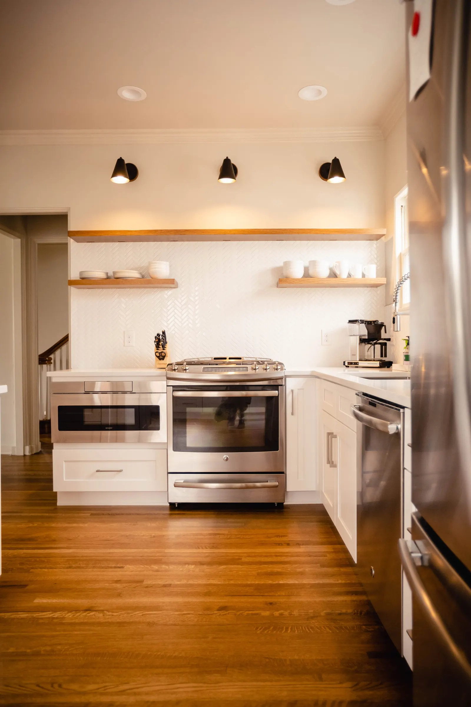 Modern kitchen with white cabinets, stainless steel appliances, wooden floating shelves with dishes, dark hardwood floors, and black ceiling lights.