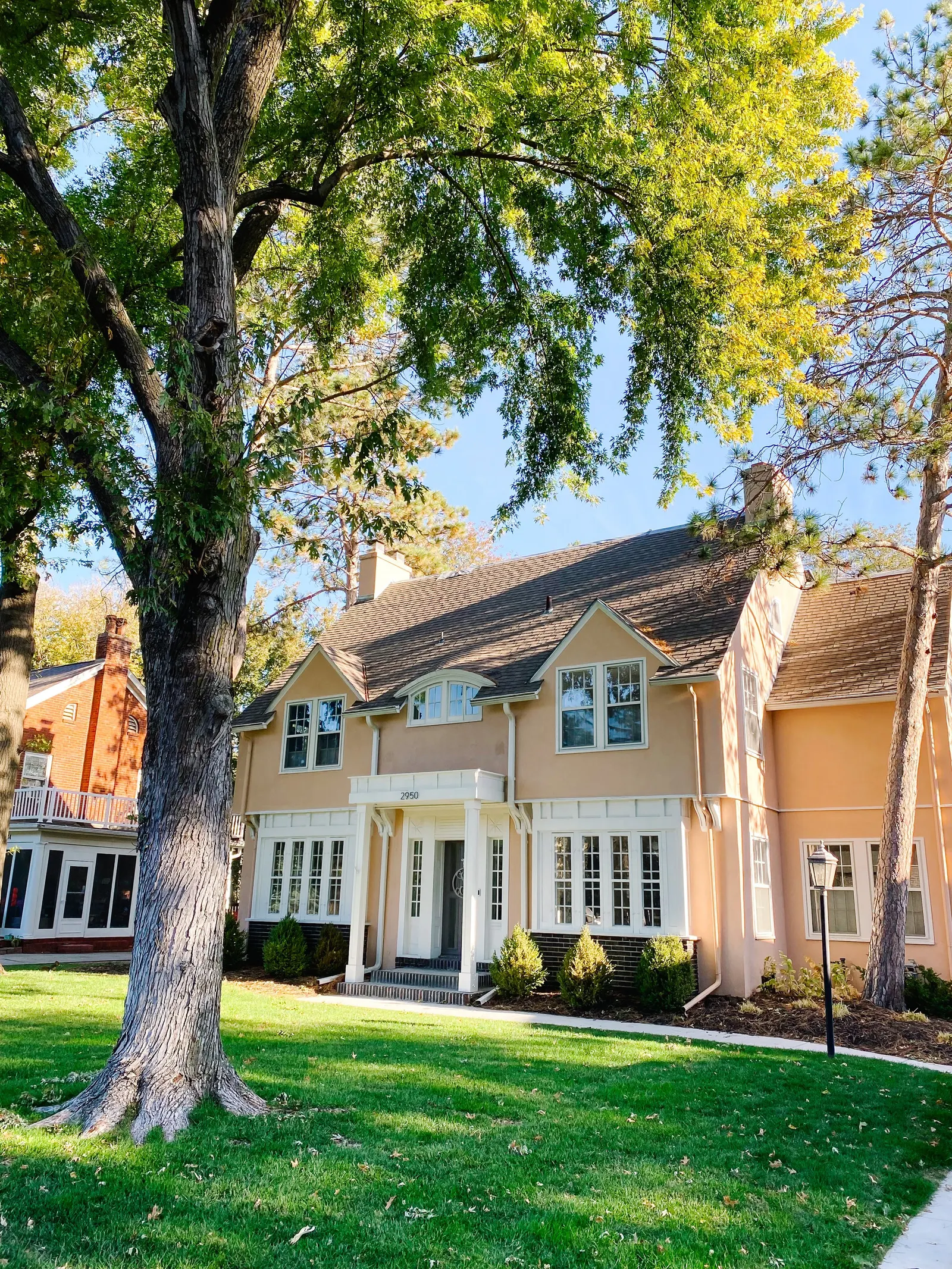 A large house with beige walls and a dark roof, surrounded by green trees and a lawn, with a sidewalk in front. Tile Roofing.