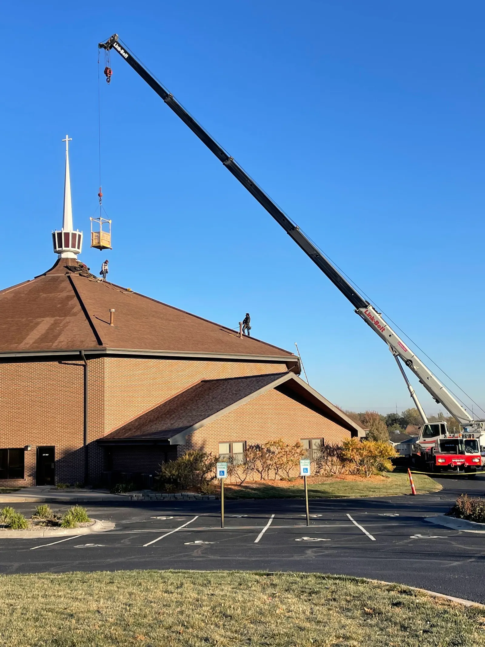 A crane is lifting a cage on top of a church steeple while workers are on the roof of the church building. Commercial Roofing.