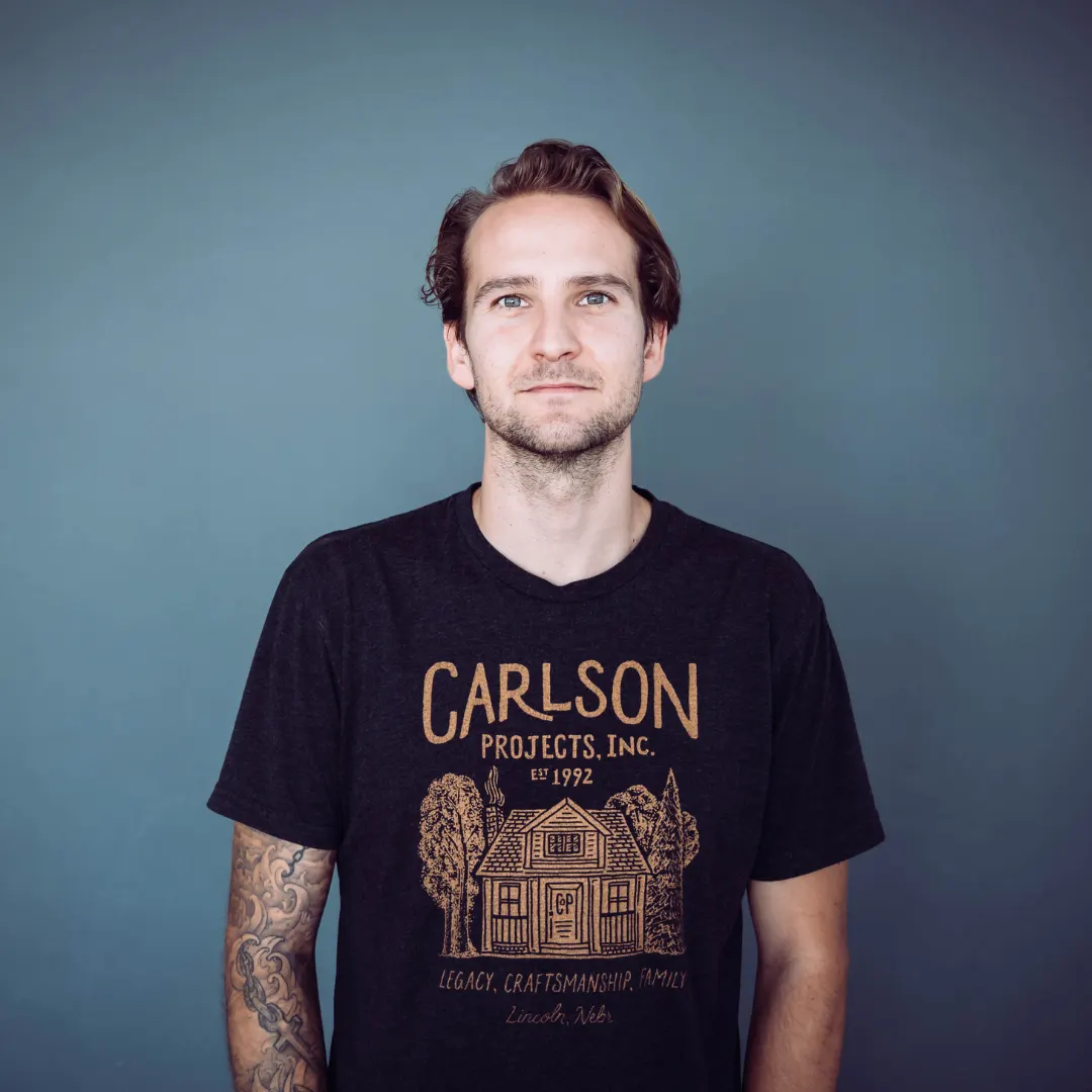 A young man with brown hair and a short beard is standing against a plain blue background. He is wearing a black T-shirt with gold-colored text and illustrations related to Carlson Projects, Inc., established in 1992, with themes of legacy, craftsmanship, and family.
