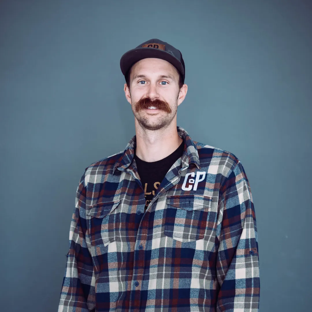 Man with a brown mustache, wearing a black and gray baseball cap, plaid shirt, and black T-shirt, smiling portraits against a teal background.