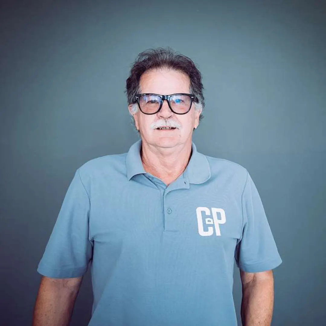 Older man with gray hair and mustache wearing glasses and a light blue polo shirt with 'CP' on the chest, standing against a dark gray background.