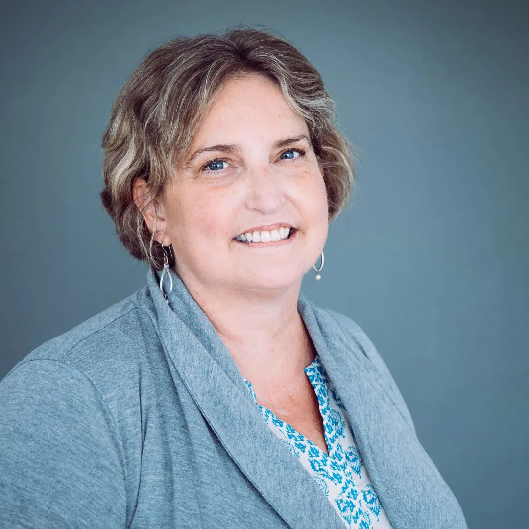 Headshot of a middle-aged woman with short, wavy blond hair, blue eyes, smiling, wearing a gray blazer over a patterned blue and white blouse, against a plain gray background.