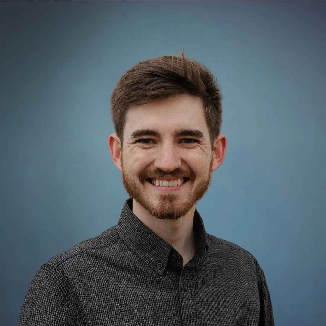 Headshot of smiling young man with brown hair, beard, wearing a black button-up shirt against a blue background.