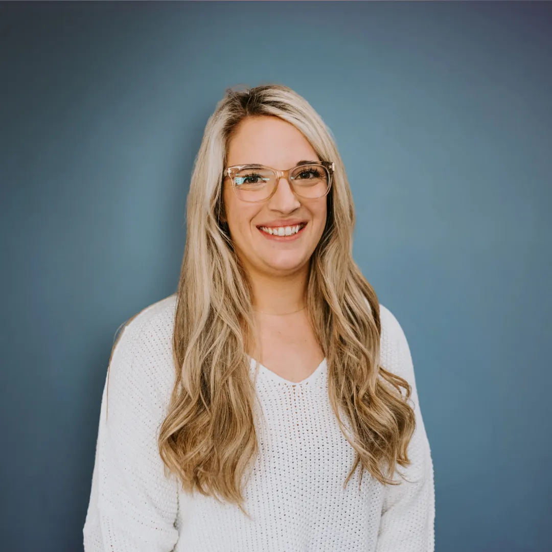 A smiling woman with long wavy blonde hair, wearing glasses and a white top, standing in front of a blue background. Social Media Coordinator and Design and Selections Support, Tessa.